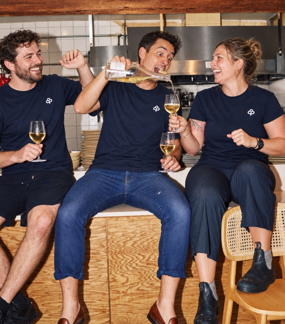 Tres miembros del equipo de un restaurante con camisetas azul marino de PAMUUC, riendo en la cocina